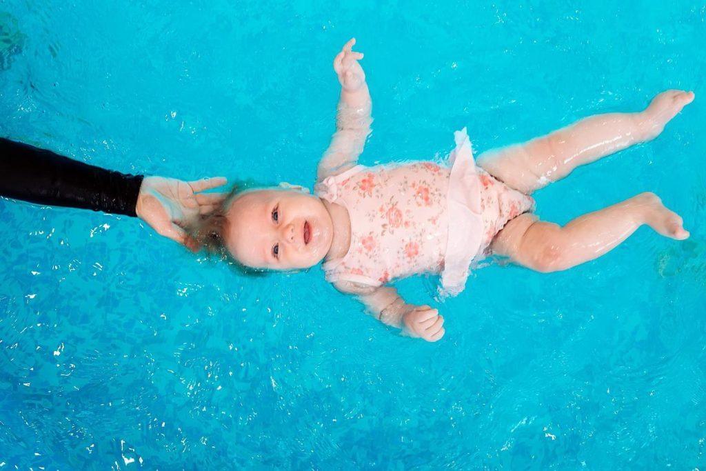 A baby wearing a floral swimsuit floats on their back in a bright blue pool. A person's hand gently supports the baby’s head.