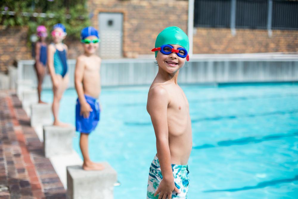 A smiling child wearing a green swim cap and red goggles stands at the edge of a swimming pool. Other children in colorful swim gear are lined up behind them, preparing to jump into the water.