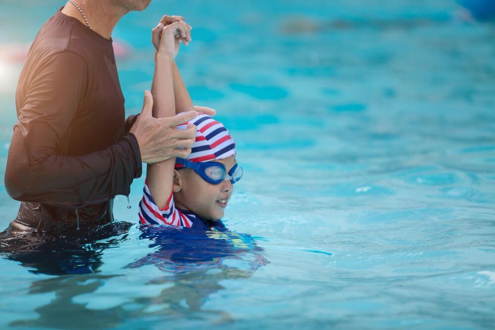 A swim instructor helps a child in a pool. The child, wearing a striped swim cap and goggles, is practicing a swimming stroke. The vivid blue water surrounds them under a sunny sky.