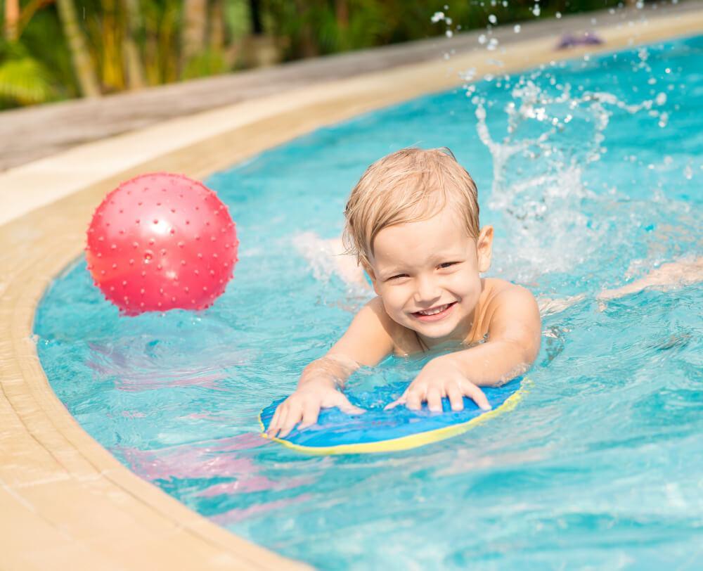 A young child with blonde hair is swimming in a pool, smiling while holding a blue and yellow kickboard. There’s a red inflatable ball floating nearby. The pool edge and some greenery are visible in the background.