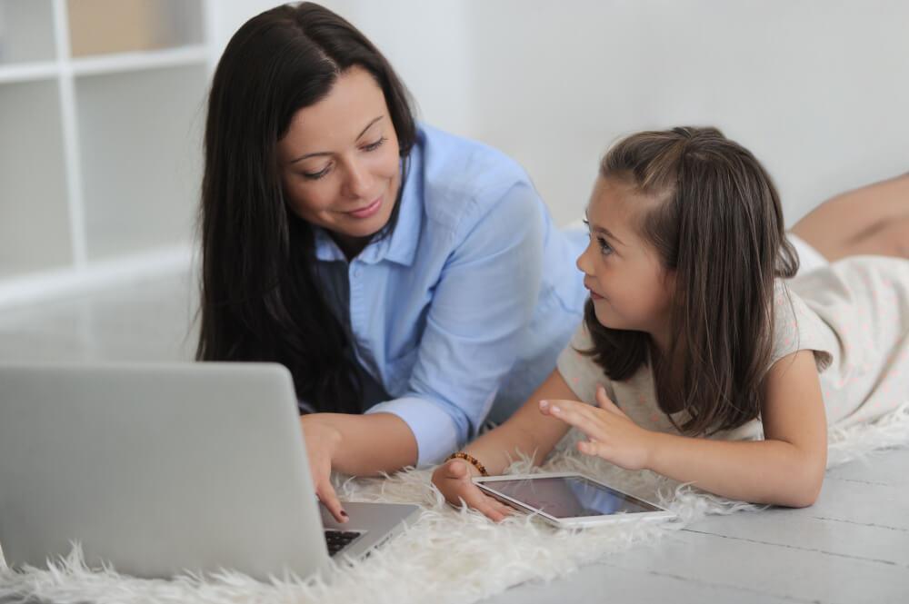 A woman and a young girl are lying on a fluffy rug, using a laptop and a tablet together. The woman is wearing a blue shirt, and the girl is in a light-colored dress. They appear engaged and happy in their interaction.