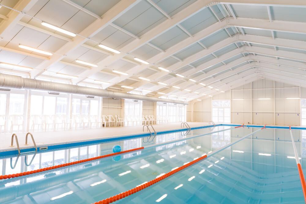 An indoor swimming pool with clear blue water and three swimming lanes marked by orange lane dividers. The facility has a high, white ceiling with bright lighting, and white chairs line the far side of the pool.