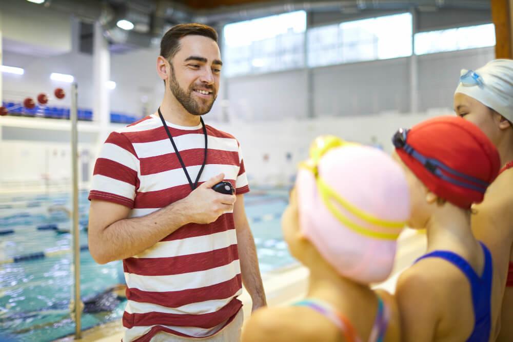 A swim coach in a striped shirt holds a stopwatch and talks to three young swimmers wearing swim caps and goggles by an indoor pool. The swimmers listen attentively, standing near the water.