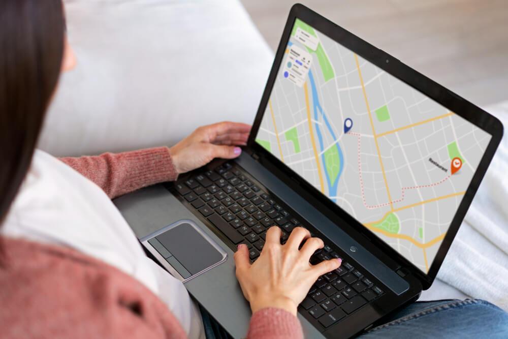 A person wearing a pink sweater is sitting with a laptop on their lap. A map with a highlighted route and two location pins is displayed on the screen.