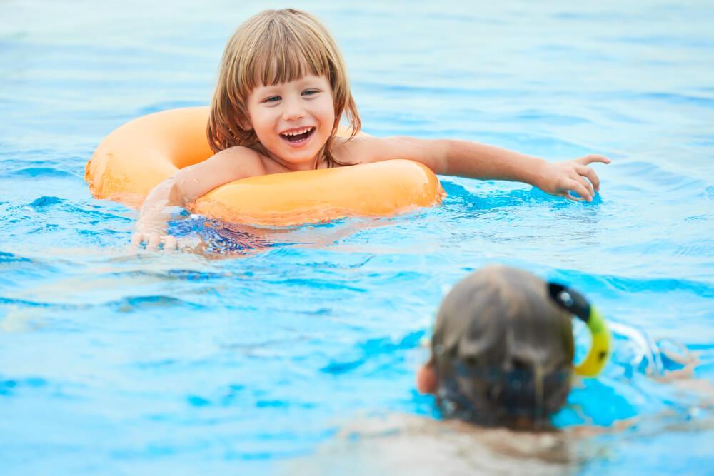 A young child with a brown bob haircut is smiling while sitting in an orange float in a pool. Another child with swim goggles and a snorkel floats nearby. The water is clear and blue, creating a cheerful, sunny atmosphere.