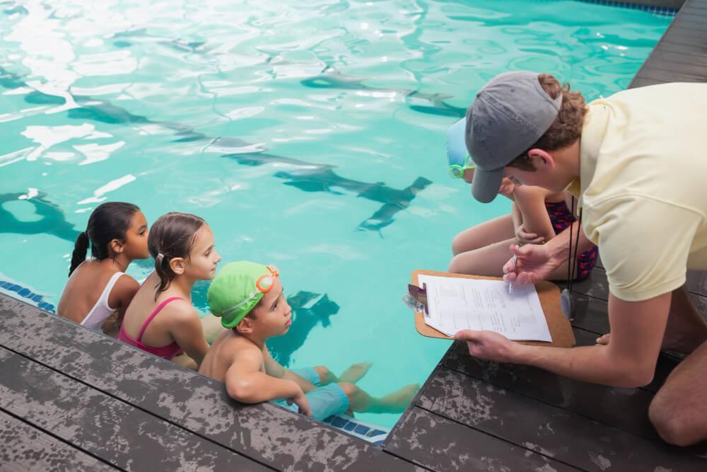 A swim instructor kneels by the pool, holding a clipboard and talking to a group of children sitting on the edge. The kids wear swimsuits and goggles, ready for their swimming lesson in the indoor pool.