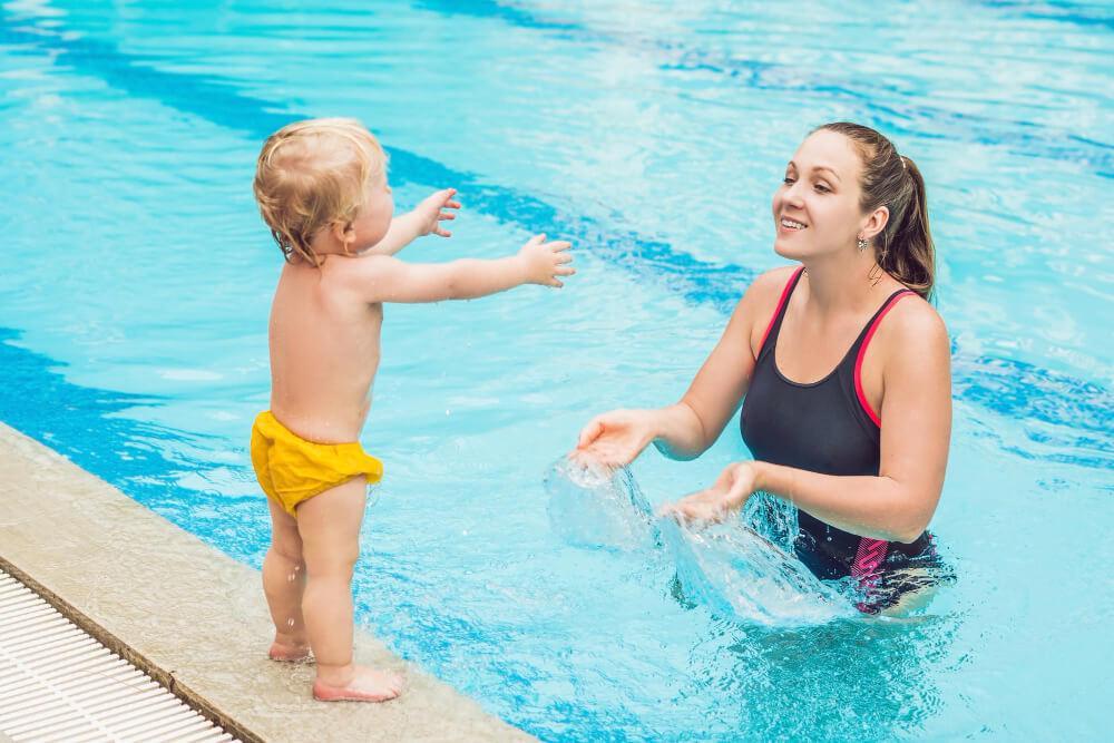 A woman in a black swimsuit stands in a pool, smiling and encouraging a toddler in yellow swim trunks on the pool's edge. The toddler has arms outstretched towards the woman.