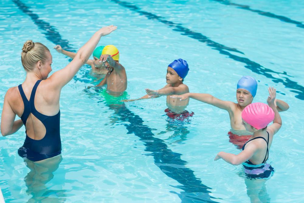 A swimming instructor in a blue swimsuit teaches four children in swim caps at a pool. The children are standing in the water, following her instructions with their arms outstretched. The pool lanes are visible.