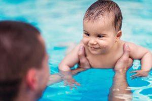 A smiling baby is held in a swimming pool by an adult as they both enjoy the moment—an early step to help little ones learn how to swim.