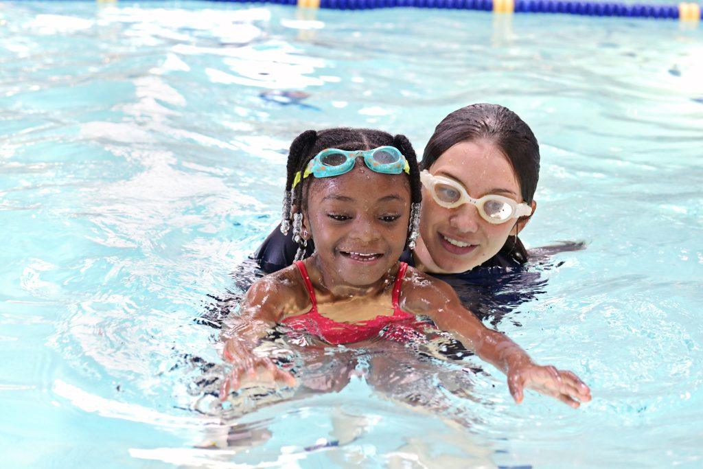 A young girl wearing swim goggles and a red swimsuit smiles while learning to swim with the help of an instructor in a swimming pool. The instructor, also wearing goggles, supports her from behind.