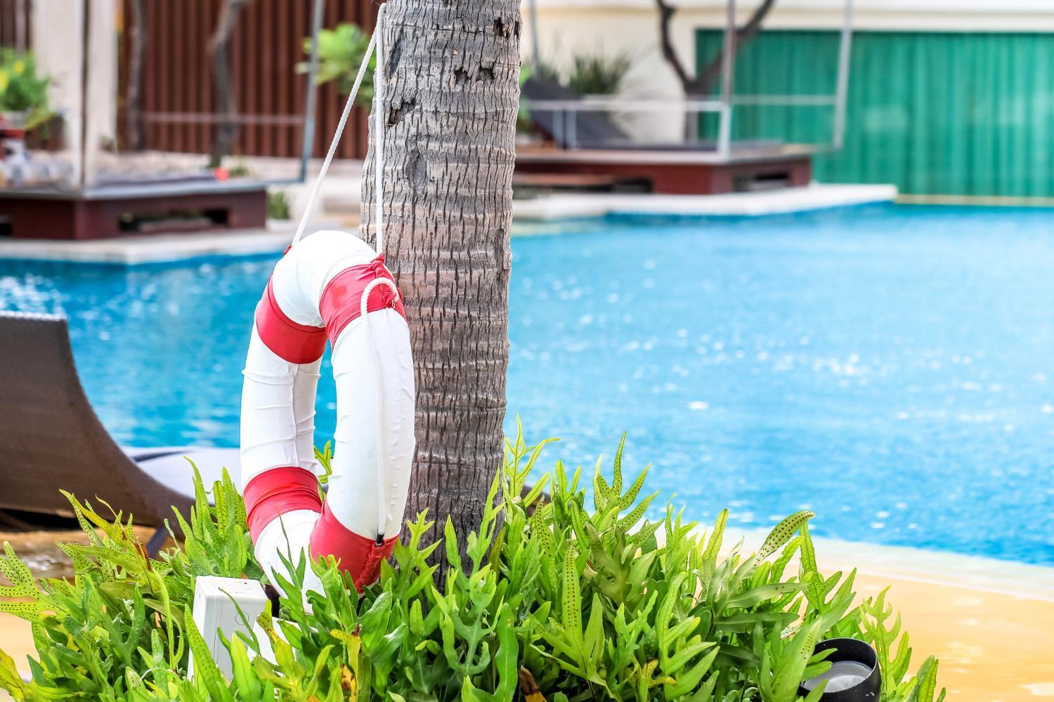 A white and red lifebuoy hangs on a palm tree near a swimming pool, surrounded by green plants, with lounge chairs and poolside structures visible in the background.