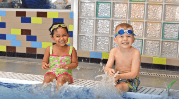 Two young children wearing swimsuits and goggles sit at the edge of an indoor pool, smiling and splashing water with their feet. The background features colorful tiles and glass blocks.