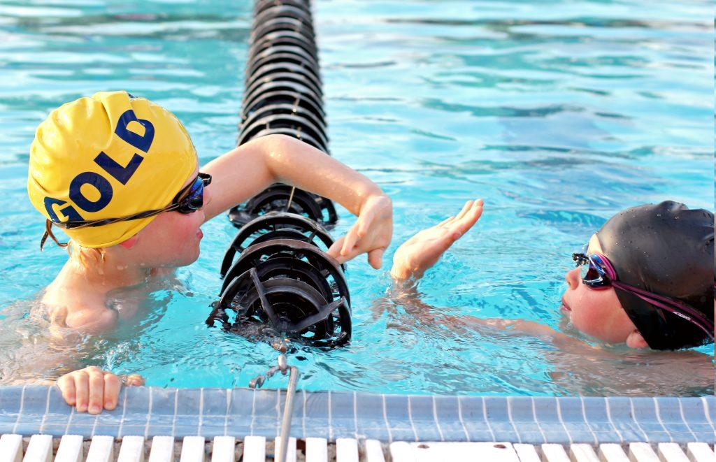 Two young swimmers, wearing goggles and swim caps, give each other a high five at the edge of a pool. One cap is yellow and says "GOLD," while the other is black. They are separated by a lane divider.