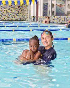 A swim instructor wearing goggles holds a smiling child in a swimming pool. Yellow and blue lane dividers and flags are visible, with other swimmers and colorful tiled walls in the background.