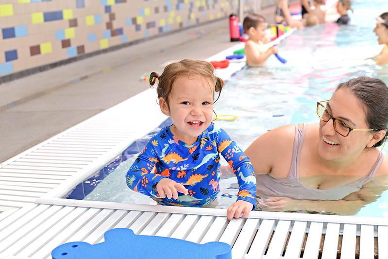 A smiling toddler in a blue swimsuit with colorful patterns stands at the edge of an indoor pool, next to an adult woman in the water—capturing a fun moment often seen at swimming schools. Other people are swimming in the background.