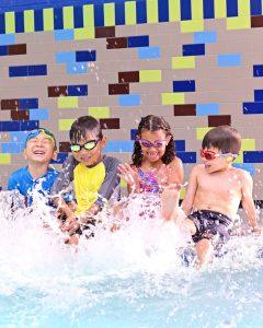 Four young children in swimsuits and goggles sit by a pool, laughing and splashing water with their feet. A colorful tile wall is in the background. The scene is joyful and playful.