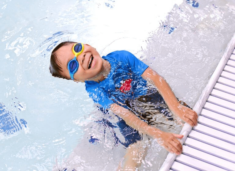 A smiling young boy wearing swim goggles and a blue shirt holds onto the side of a pool, partially submerged in the water.