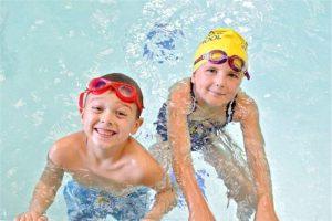 Two children in a swimming pool smile up at the camera. One wears red goggles and the other wears a yellow swim cap and purple goggles. Both are holding onto the pool edge.